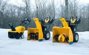 Three stages and sizes of snow blowers by Cub Cadet are displayed on a icy parking lot with leafless trees in the background